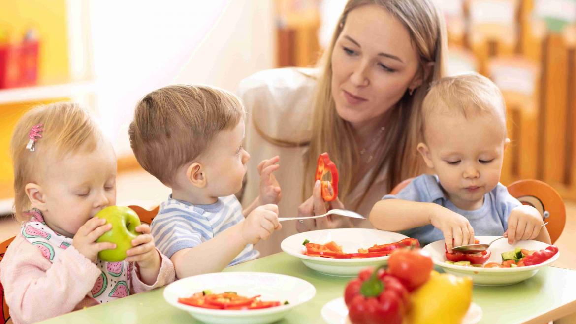 Kinder essen am Tisch in einer Kita mit Erzieherin