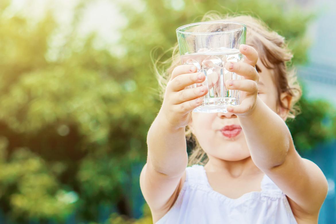 Kleines Mädchen hält ein leeres Wasserglas in die Höhe. Sein Gesicht ist durch das Glas verdeckt.